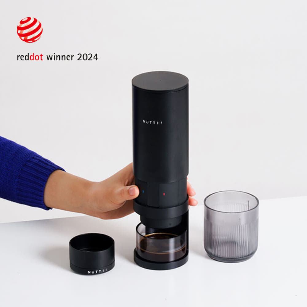 Black coffee maker with a glass of coffee on a white background, featuring the Red Dot Award logo.